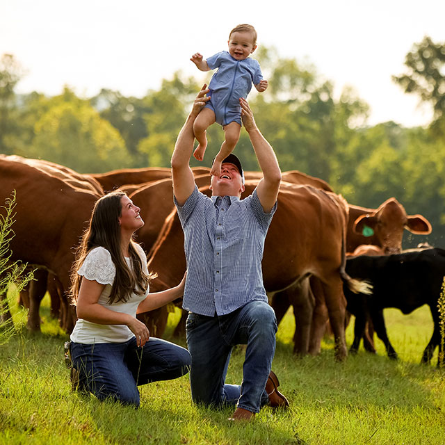 A husband and wife pose, holding their son in front of a herd of red cattle.